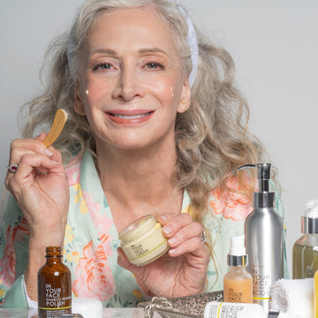 5 REASONS TO SWITCH TO NATURAL SKINCARE: a mature, pretty woman applying IN YOUR FACE CREAM and smiling, surrounded by bottles of skincare products