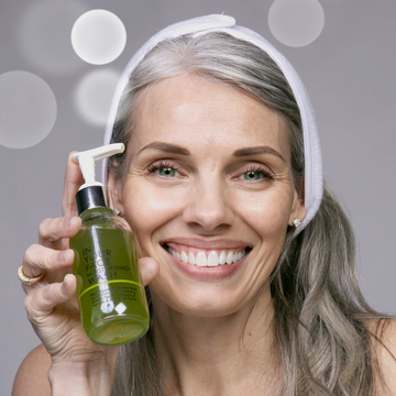 a pretty, mature woman with long grey hair holding IN YOUR FACE MOISTURIZING WASH to her cheek and smiling