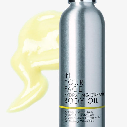 Silver bottle of 'In Your Face Hydrating Creamy Body Oil' with yellow oil smear on white background
