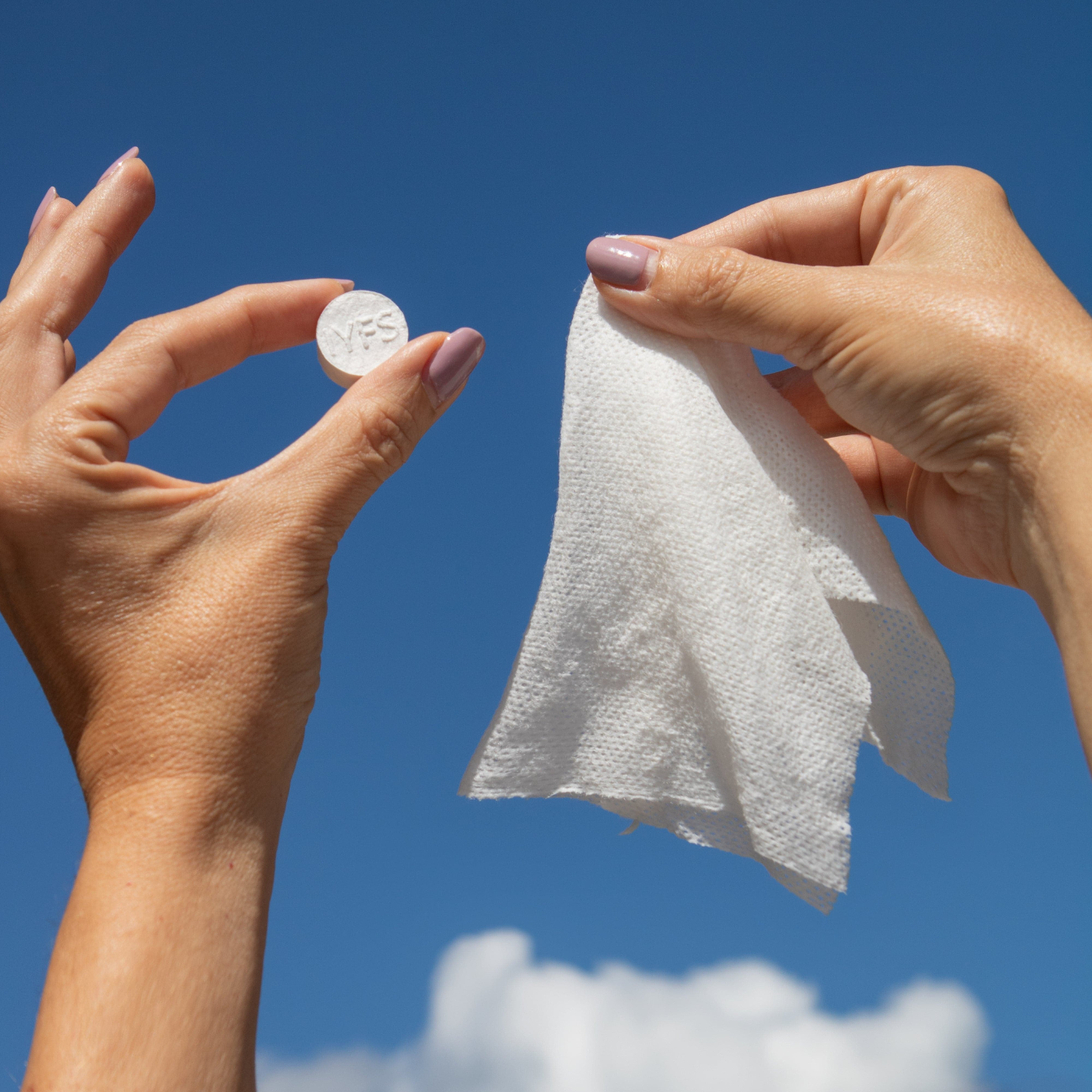 A photo of 2 hands against the sky. One is holding a cleansing cloth that hasn't been soaked in water and is the round shape. The next is holding a cloth that has been expanded in water and is a square shape.