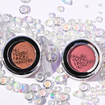 A photo of the bronzy brown-colored IN YOUR FACE SHIMMER next to the IN YOUR FACE COLOR (which is a pretty rose tone), surrounded by iridescent sparkly gems. They are reflecting light.