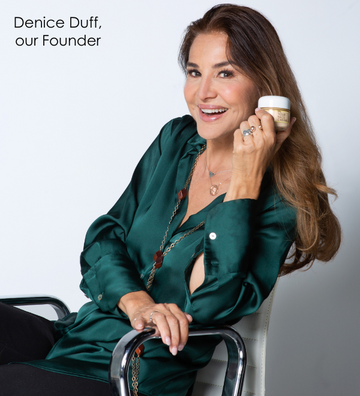 Denice duff, our founder, sitting on a chair smiling, wearing a silky green button down and holding up our hero product THE CREAM
