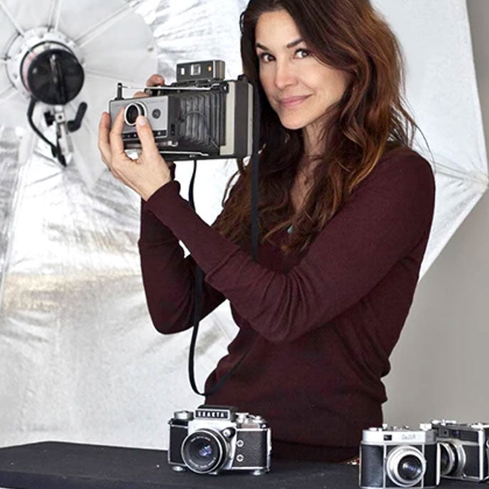 denice duff holding a camera up near her face with photography equipment in the background. She looks professional and is wearing a long sleeved burgundy top