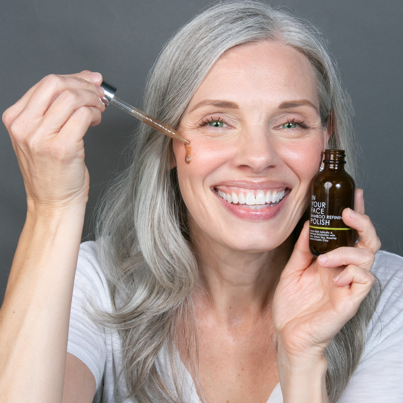 an image of a smiling, pretty, early 50s woman with long grey hair holding the BAMBOO POLISH in her hand and a pipette of it to her cheek.