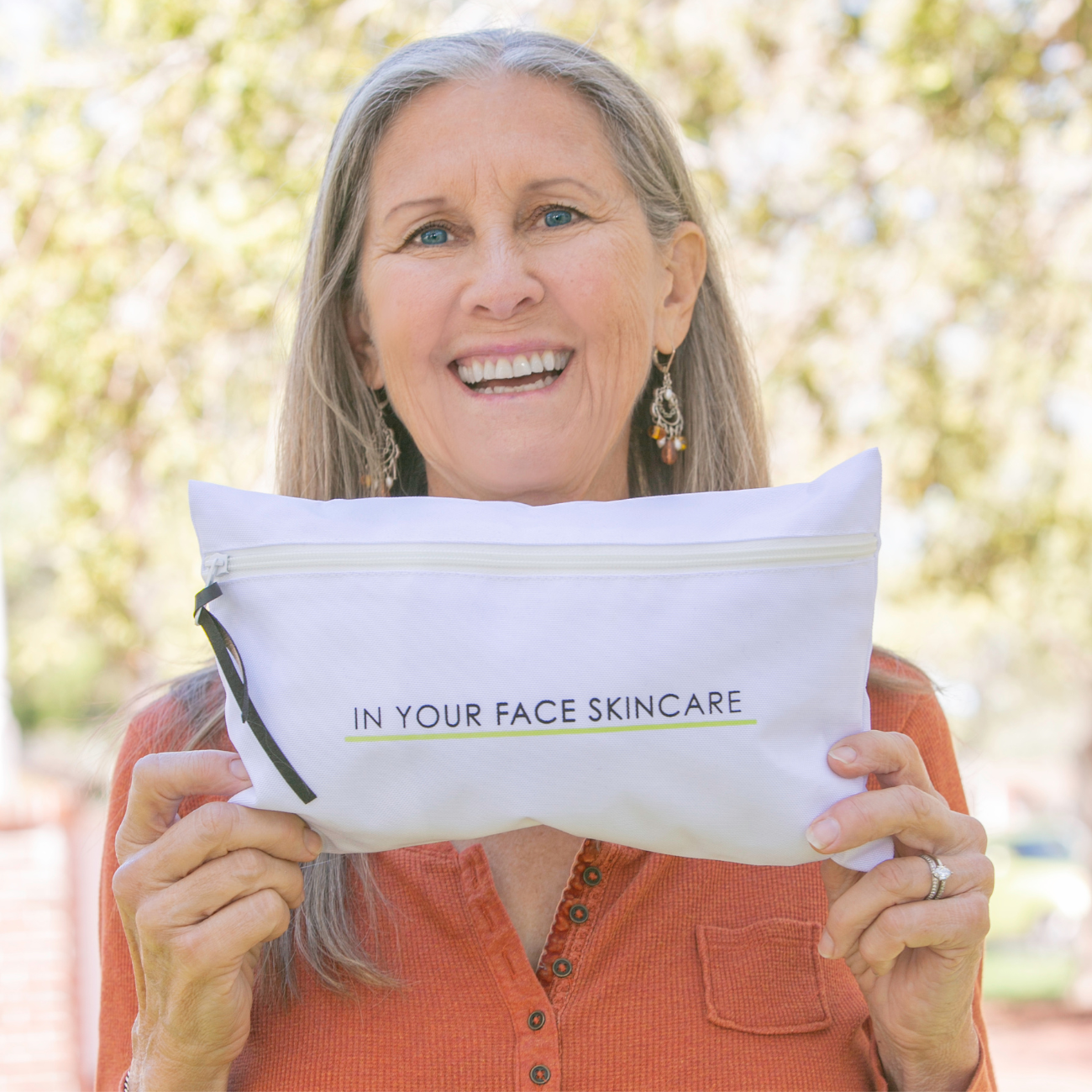 an image of a white rectangular zippered makeup bag that says "IN YOUR FACE SKINCARE" being held by a mature women with long grey hair wearing a rust-colored shirt on a background of trees