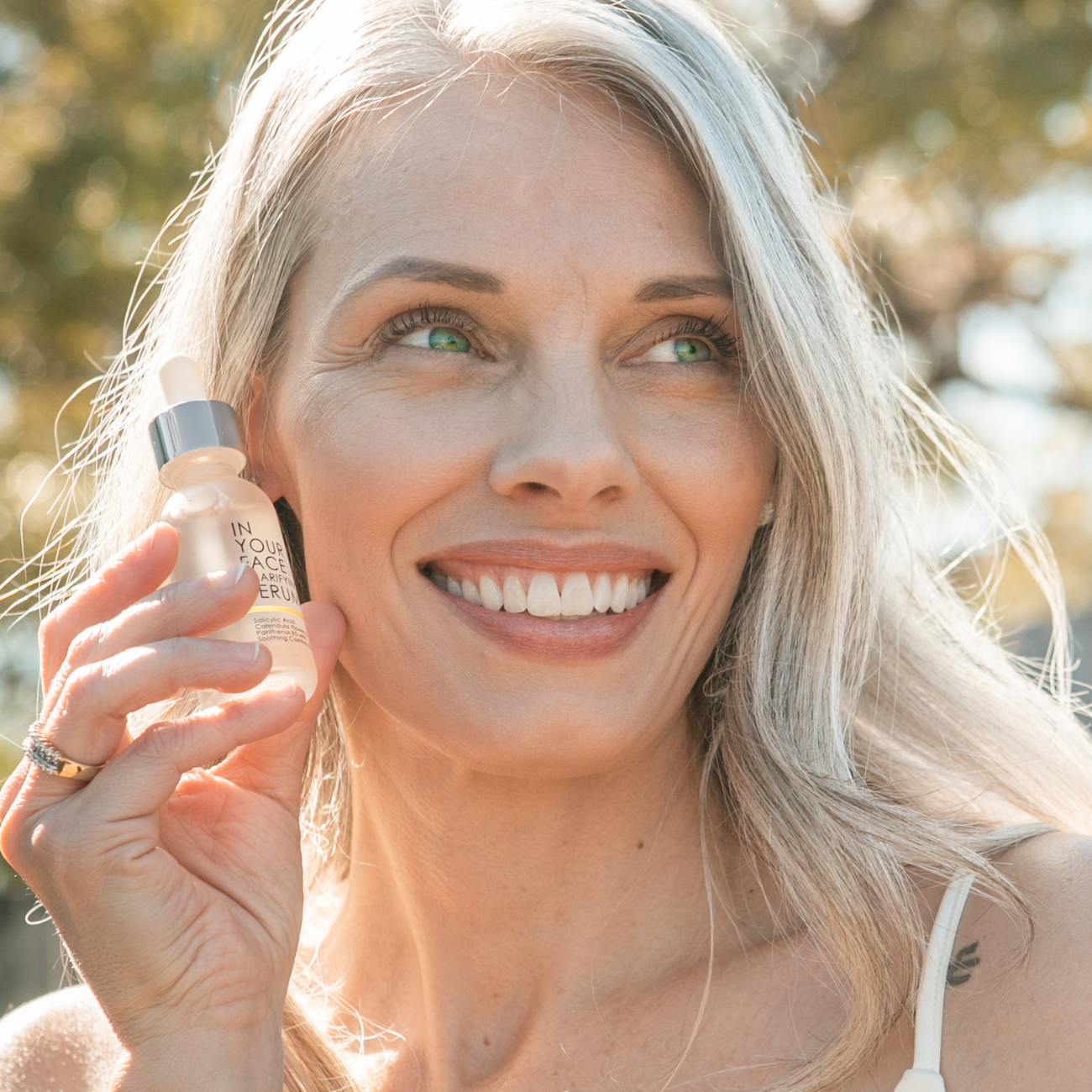 Image of smiling grey-haired woman holding bottle of CLARIFYING SERUM.