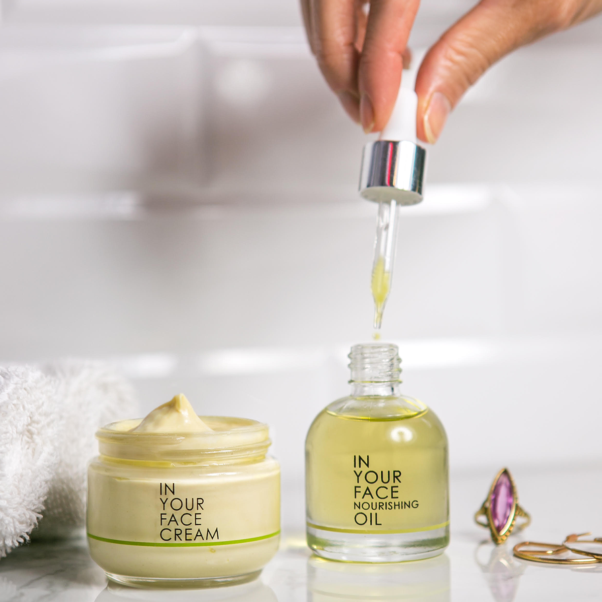 an image of the IN YOUR FACE CREAM and IN YOUR NOURISHING OIL on a tiled background. A hand is reaching down to the OIL's pippette and taking some oil out of the bottle. 