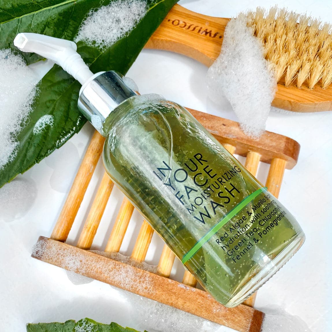 A photo of the MOISTURIZING WASH laying on a wooden soap dish next to a couple leaves and a body brush and surrounded by foam bubbles.