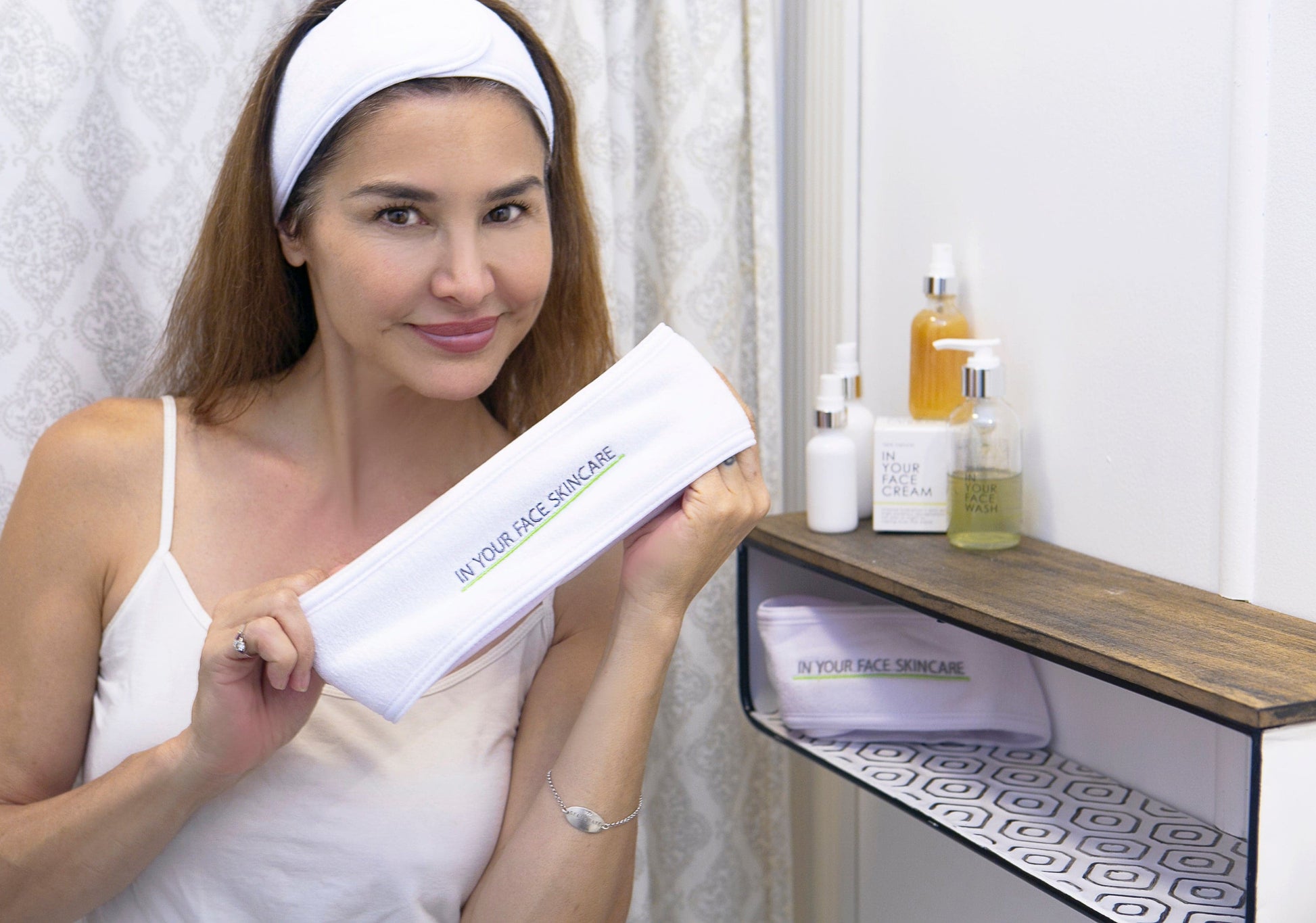 an image of Denice Duff in a bathroom wearing an IN YOUR FACE SKINCARE HAIR WRAP and holding one in her hands. She is next to a shelf with a few skincare products on top.