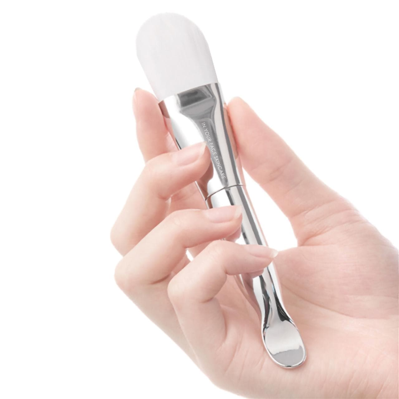 an image of a hand holding the IN YOUR FACE MASK BRUSH on a white background. It's a silver color with white bristles.