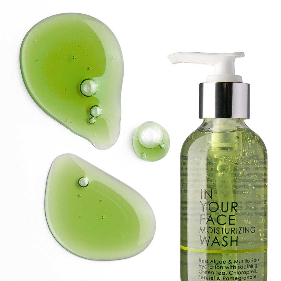 A photo of the IN YOUR FACE MOISTURIZING WASH on a white background. The bottle shows the wash inside to be a medium green color. The bottle also says "Red Algae & Murillo Bark hydration with soothing Green Tea, Chlorophyll, Fennel & Pomegranate". Next to the bottle it shows 2 big dollops and a small dollop of the wash showing the green color.