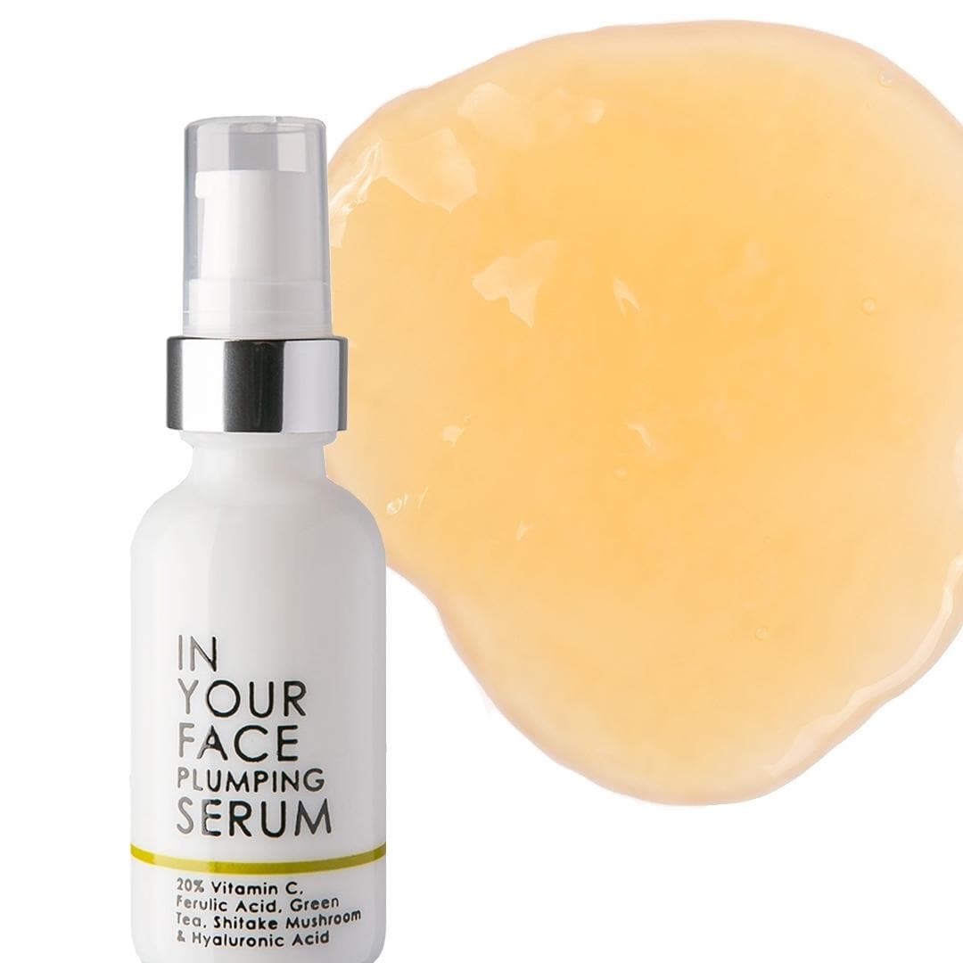 a clean image of our vitamin c plumping serum on a white background with a small shadow. The bottle saying: IN YOUR FACE PLUMPING SESRUM, 20% Vitamin C, Ferulic Acid, Green Tea, Shiitake Mushroom and Hyaluronic Acid. Next to the bottle there is a pale orangey round smear of the liquid.