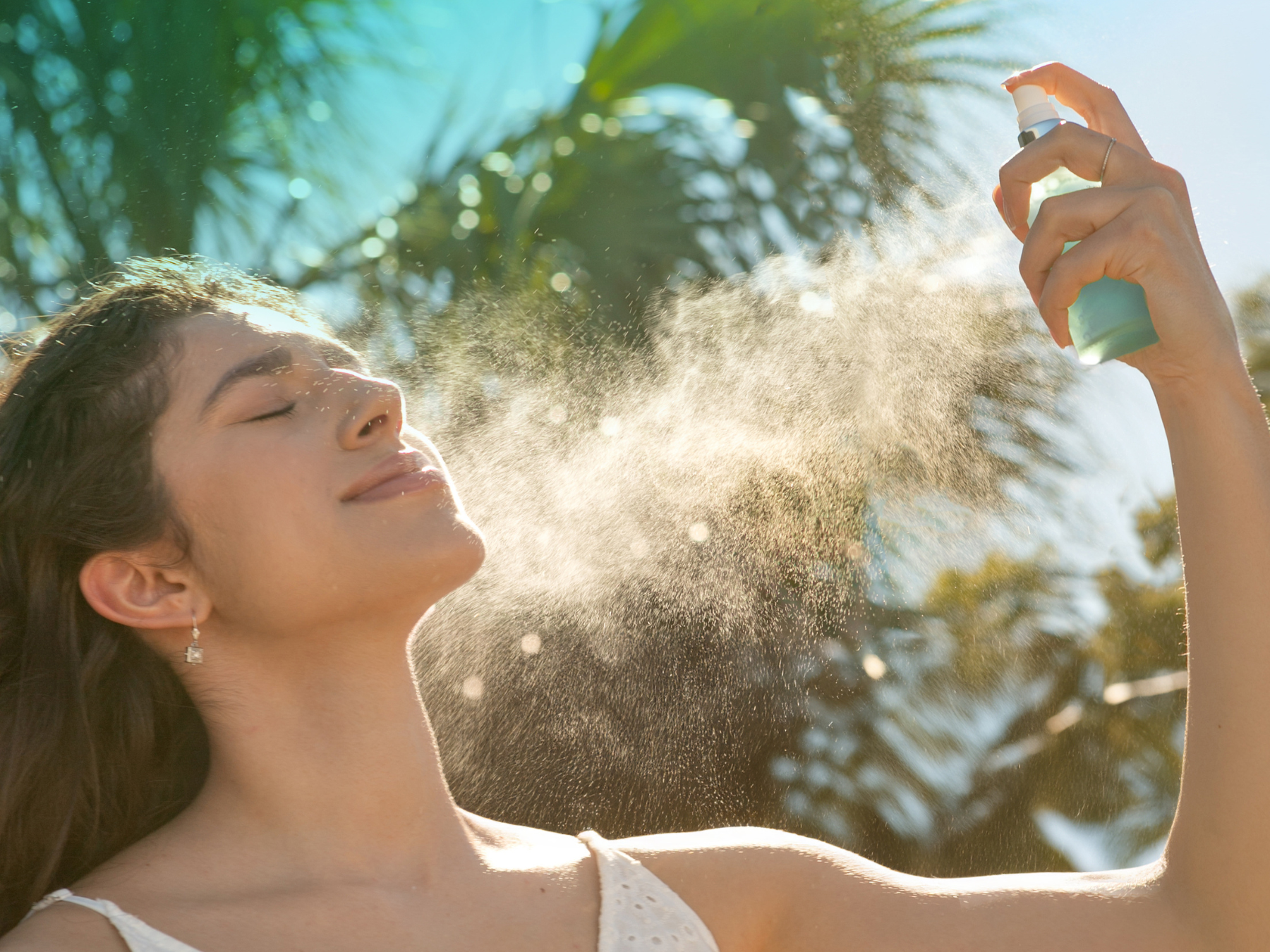 Brunette woman in front of palm tree spraying face mist 