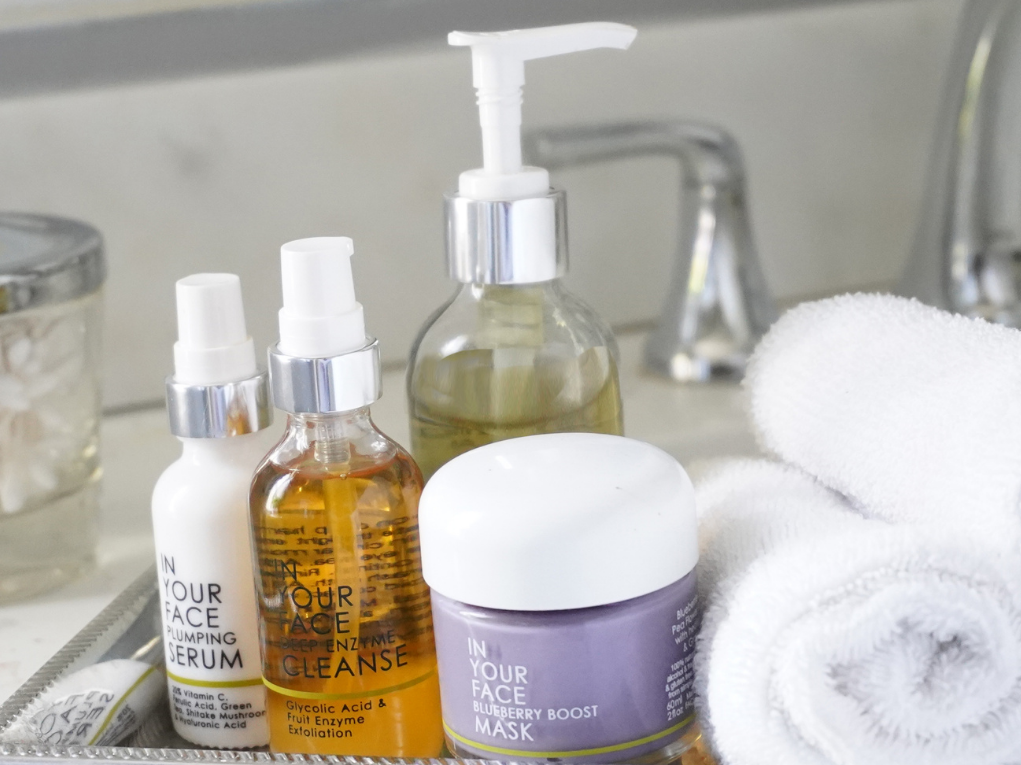 Easy Steps to a Basic Skincare Routine! A group of IN YOUR FACE SKINCARE products on a tray in a bathroom with a faucet behind them and towels beside them