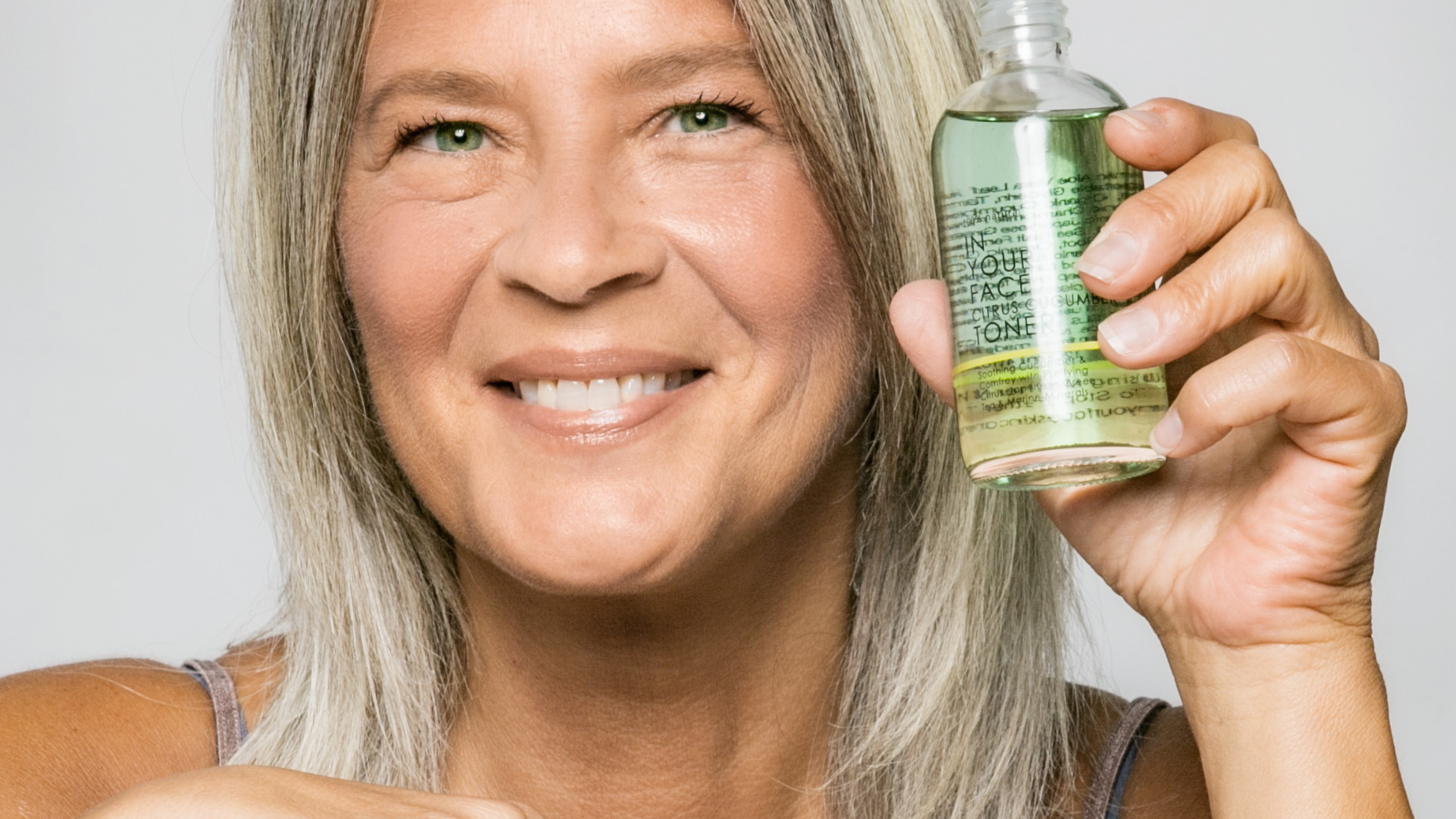 SKINCARE MYTHS: TO TONE, OR NOT TO TONE: a smiling, mature woman is holding up a bottle of IN YOUR FACE CITRUS CUCUMBER TONER
