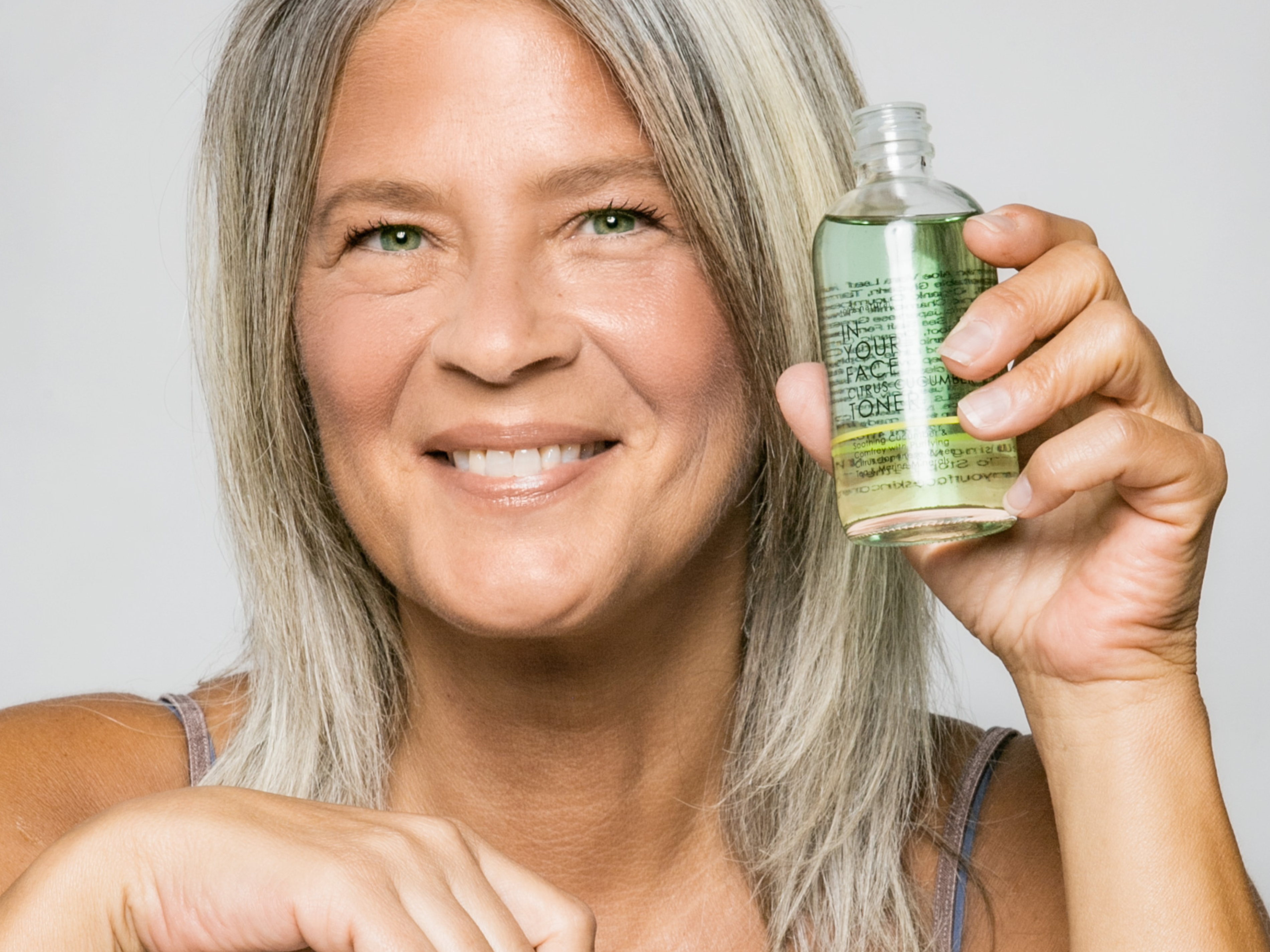 SKINCARE MYTHS: TO TONE, OR NOT TO TONE: a smiling, mature woman is holding up a bottle of IN YOUR FACE CITRUS CUCUMBER TONER