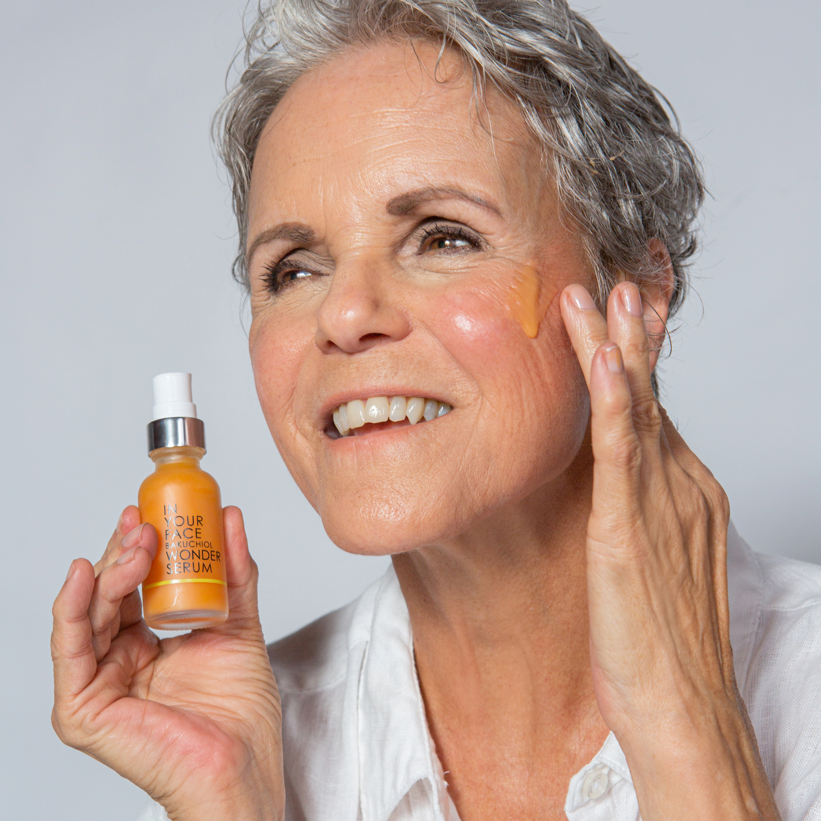 an image of a pretty, short grey-haired mature woman, holding a bottle of the BAKUCHIOL WONDER SERUM and applying some to her cheek and smiling.