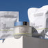 A skincare set featuring a jar of IN YOUR FACE 5X RESURFACING PEEL PADS in the center, flanked by two white pouches of the same product on a white pedestal against a blue sky background.