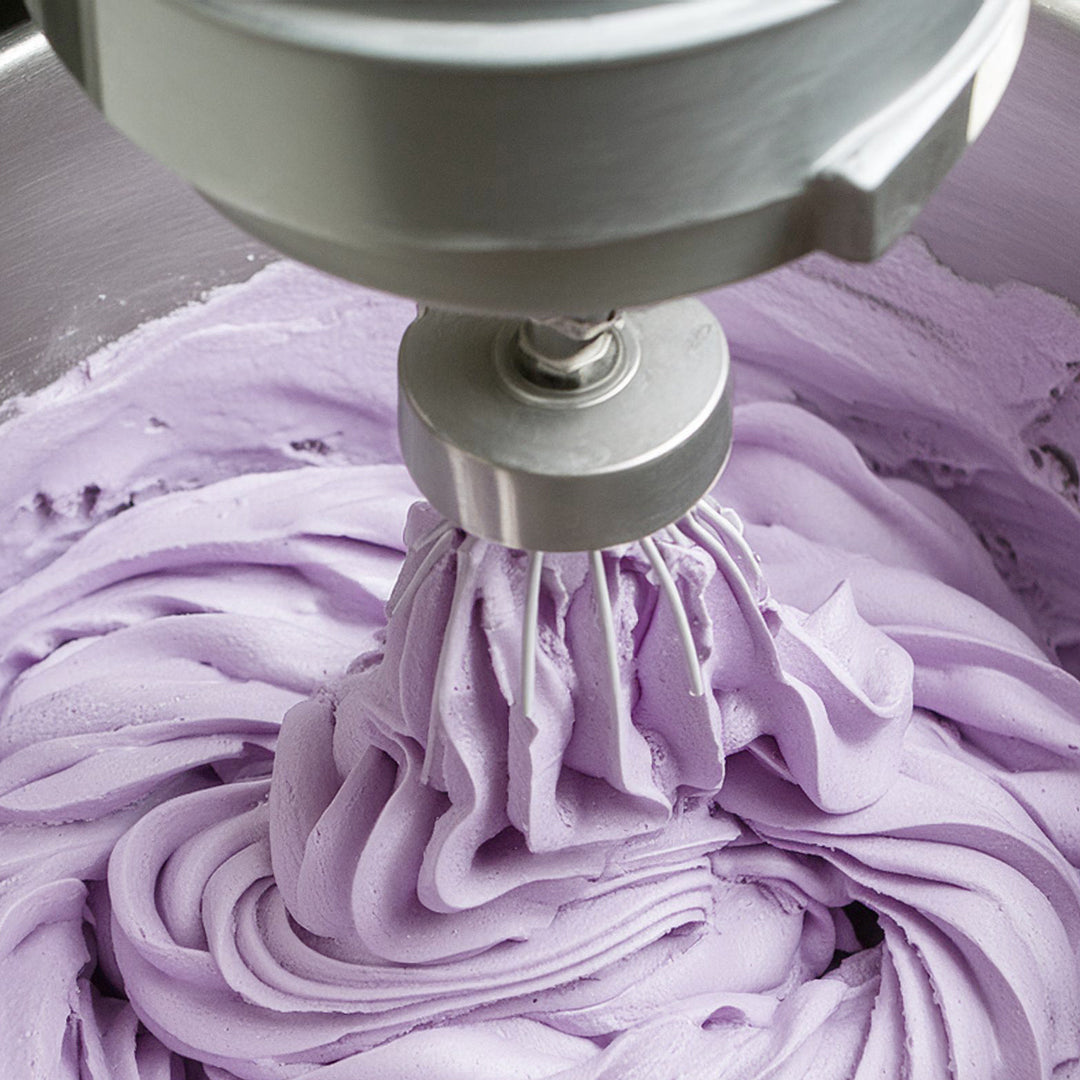 a metal bowl with a lilac-colored fluffy whipped skincare product inside, showing it being blended with a metal mechanical whisk.