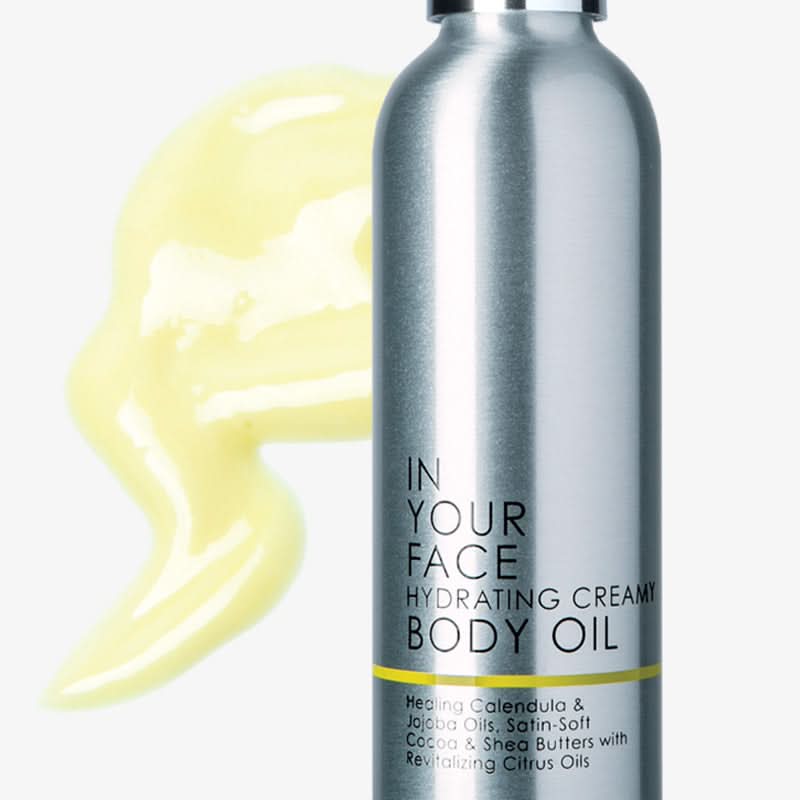 Silver bottle of 'In Your Face Hydrating Creamy Body Oil' with yellow oil smear on white background