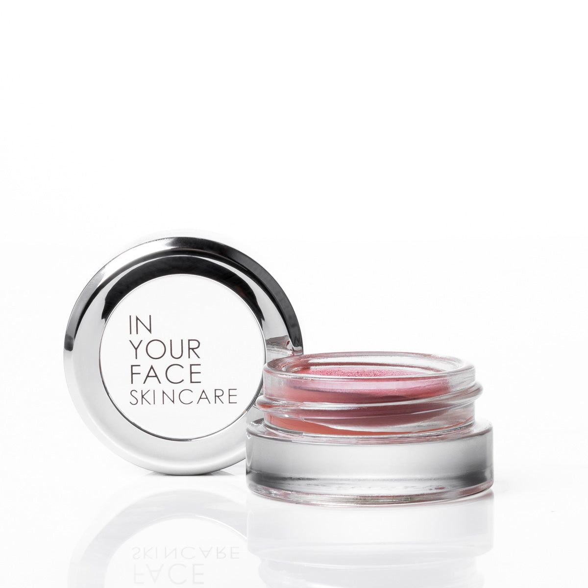 An open jar of pink cosmetic cream next to its silver lid, which displays the brand name 'IN YOUR FACE SKINCARE', on a clean white background with a soft reflection.