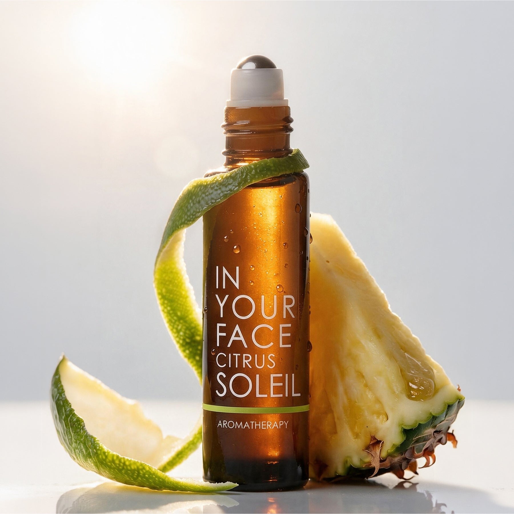 An amber glass rollerball bottle labeled 'IN YOUR FACE CITRUS SOLEIL' stands on a white surface with a citrus peel wrapped around it, flanked by pineapple slices against a bright white background.
