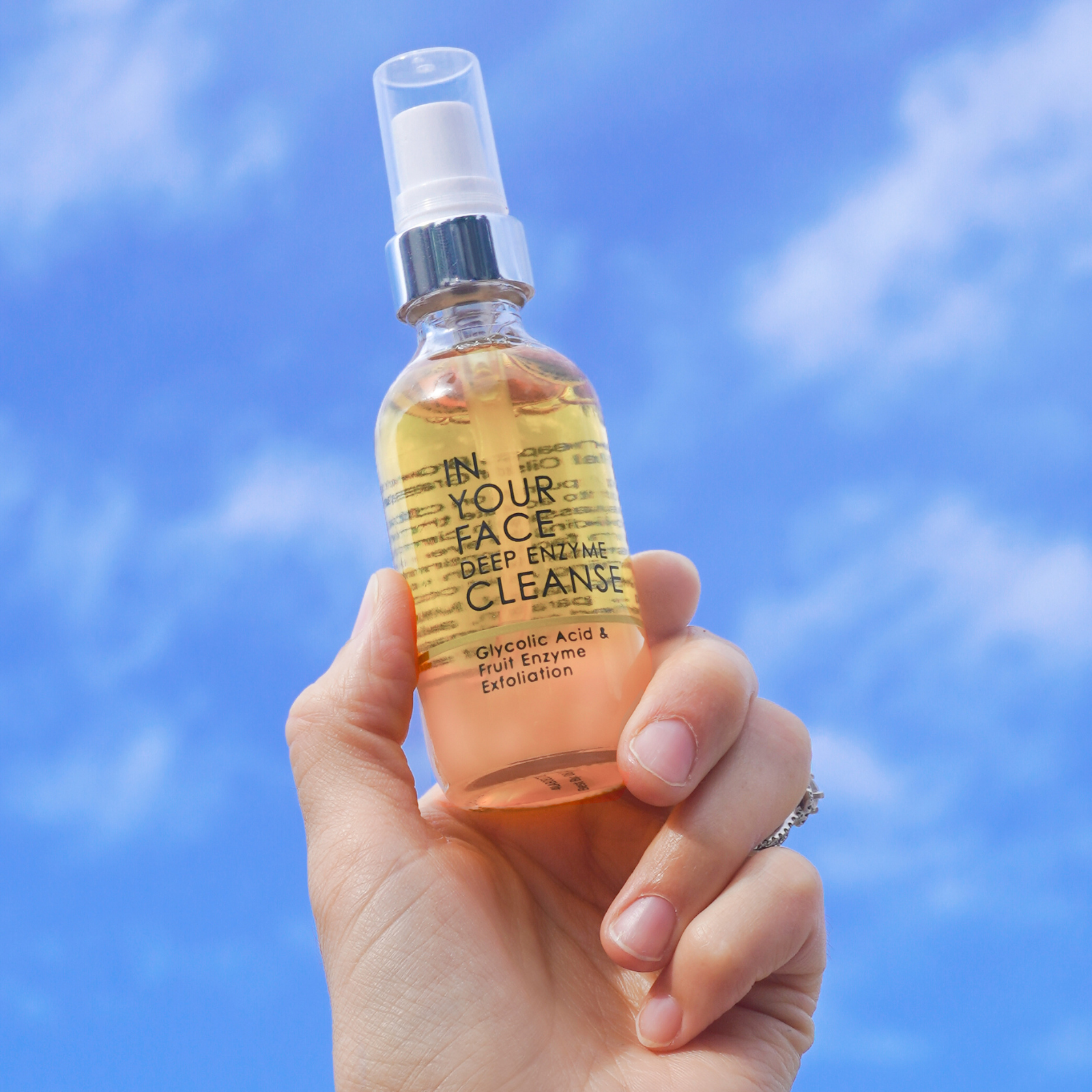 a bottle of the DEEP ENZYME CLEANSE being held in a hand up against a pretty blue sky with some out-of-focus clouds.