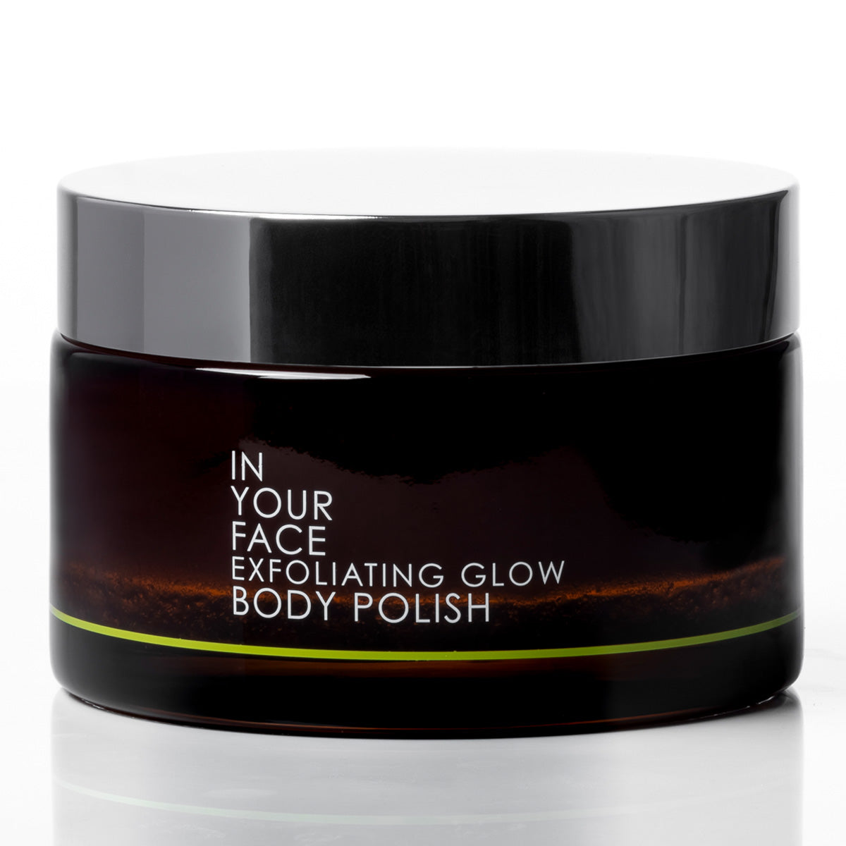 A dark amber glass jar of 'In Your Face Exfoliating Glow Body Polish' with a glossy black lid, set against a clean white background with a soft reflection.