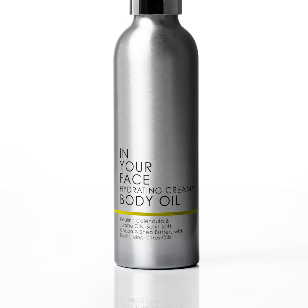 A metallic silver cylindrical bottle of body oil with a black spray top and black text on the front, photographed on a clean white background with a soft reflection below.