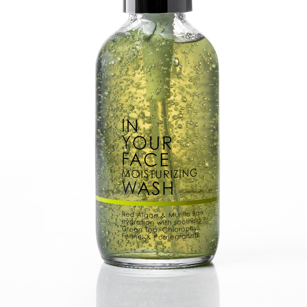 A clear glass bottle of face moisturizing wash filled with greenish-yellow liquid and visible bubbles, with a black dropper cap and black text reading 'IN YOUR FACE MOISTURIZING WASH' on a white background.