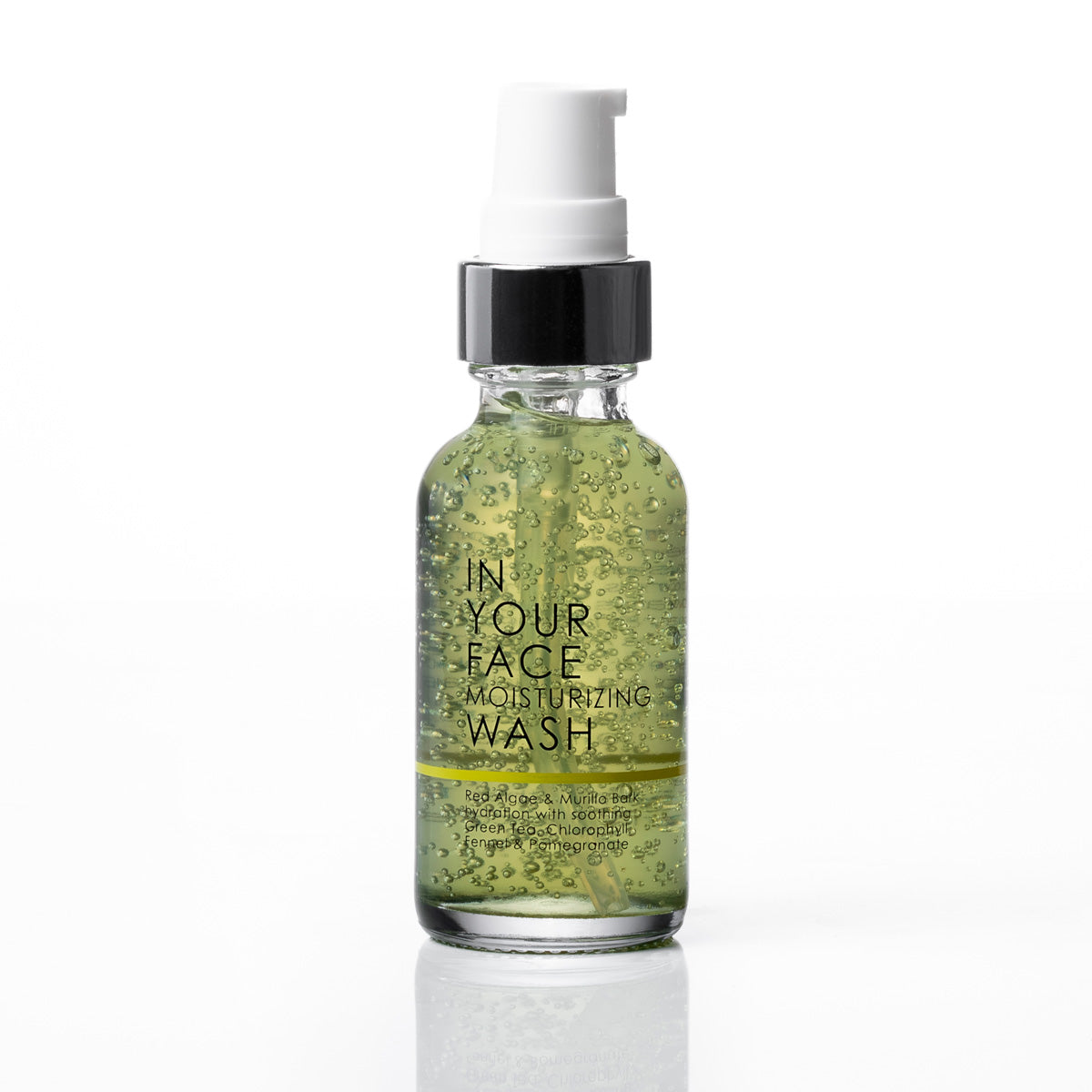 A clear glass bottle of 'In Your Face Moisturizing Wash' filled with green gel and suspended bubbles, featuring a black collar and white pump dispenser, set against a clean white background with a soft reflection.