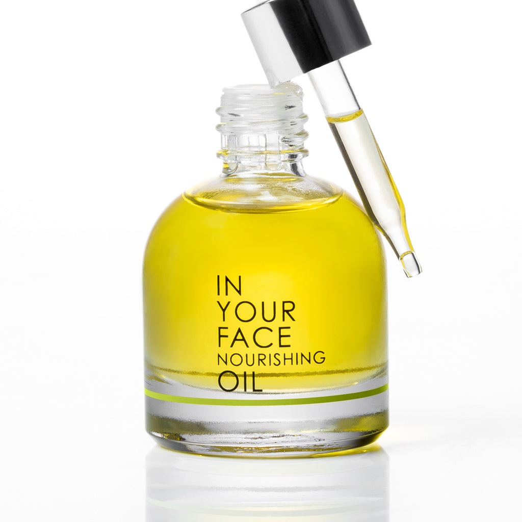 A clear glass bottle of In Your Face Nourishing Oil filled with yellow oil, shown with a black-capped dropper resting against the neck on a white background.