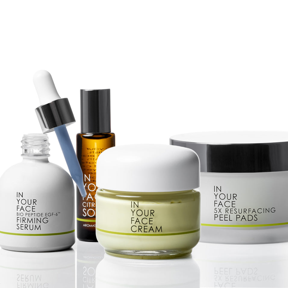 Four skincare products from IN YOUR FACE COSMETHICS arranged on a reflective white surface: a white bottle of Bio-Peptide EGF-4 Firming Serum with a blue dropper, an amber bottle of Skin-Citronessence, a clear jar of Face Cream with pale yellow cream and a white lid, and a white jar of 5X Resurfacing Peel Pads with a black lid.