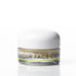 A small clear jar of face cream with a white lid and a label reading '100% Natural' and 'IN YOUR FACE CREAM', shown on a clean white background with a faint reflection below.