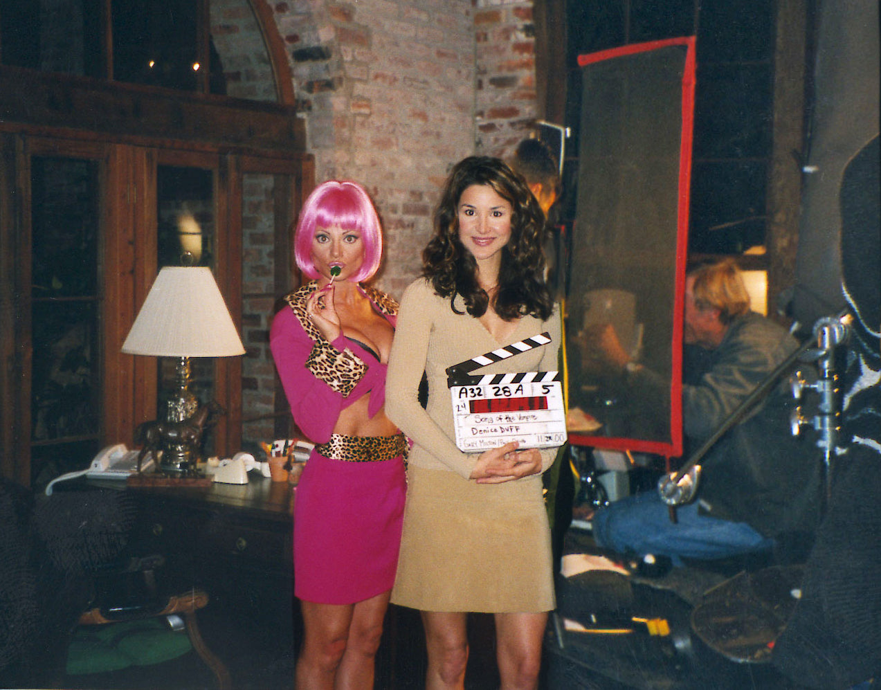 A behind-the-scenes photo of a film set featuring two women. On the left, a woman wears a bright pink wig and a magenta mini dress with gold leopard-print trim and belt.