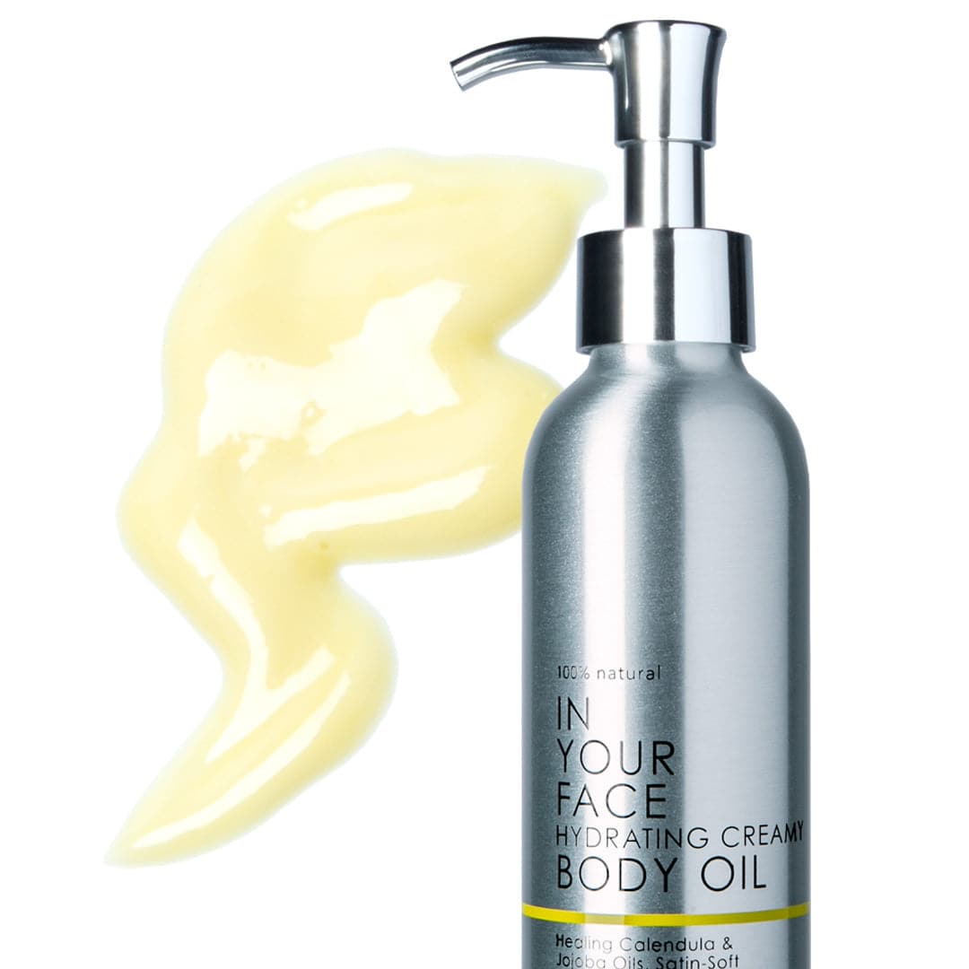 A metal bottle with a spout pump of the IN YOUR FACE HYDRATING CREAMY BODY OIL, saying "100% natural", with the words "Healing Calendula & Jojoba Oils, Satin-Soft Cocoa & Shea Butters with Revitalizing Citrus Oils", also showing a smear of the oil, and it's a light buttery yellow color.