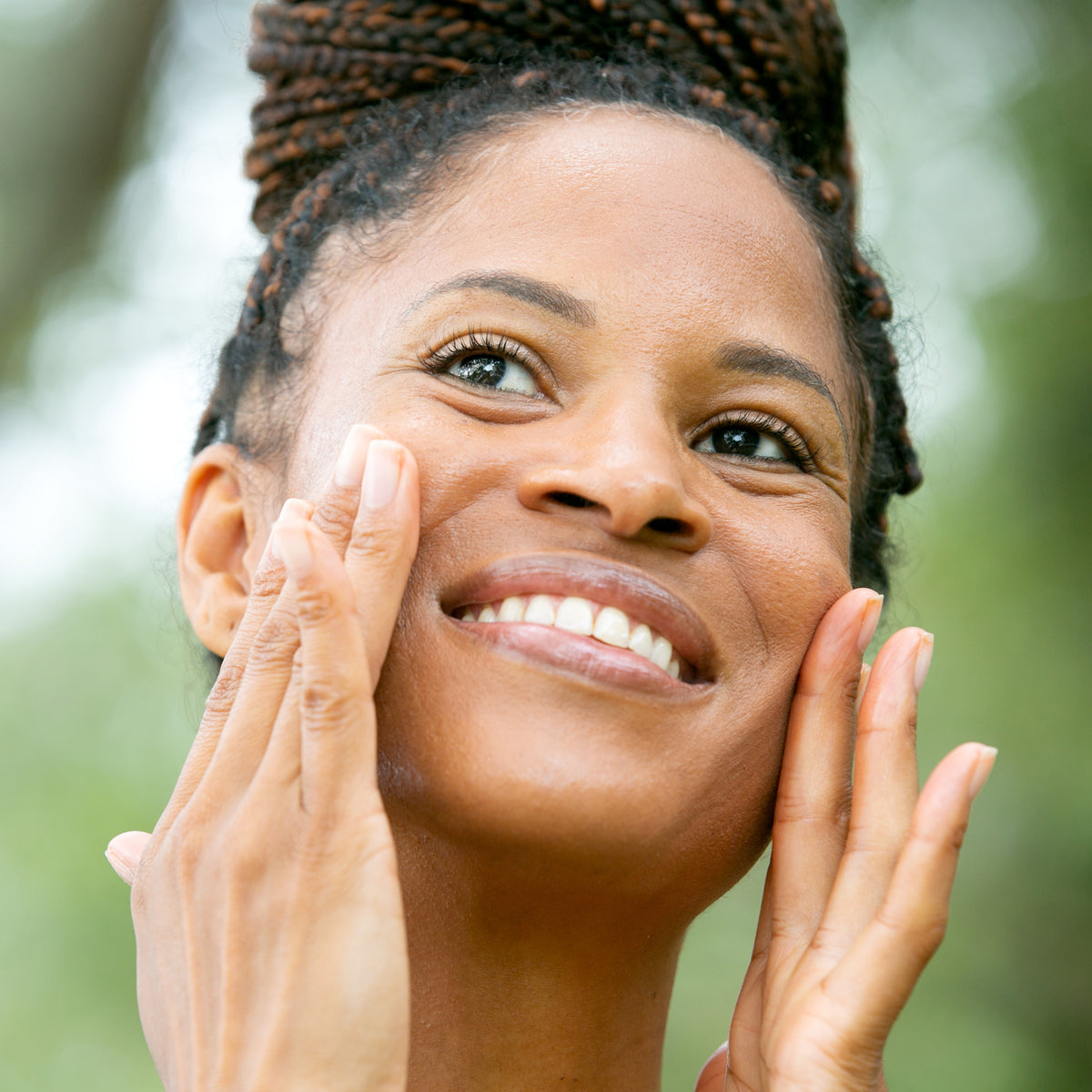 A close-up portrait of a smiling woman with braided hair, touching her face, set against a soft blurred green outdoor background.