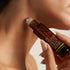 A close-up lifestyle shot of a person's neck and jawline as they apply an amber glass roll-on skincare bottle to their skin. The bottle is labeled 'YOUR FACE YOUR SOUL' and 'ACROMANCERY'.