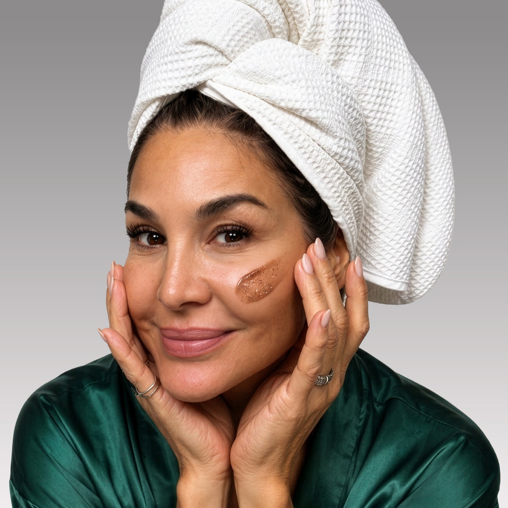 Woman applying skincare product to her face with a towel on her head against a gray background