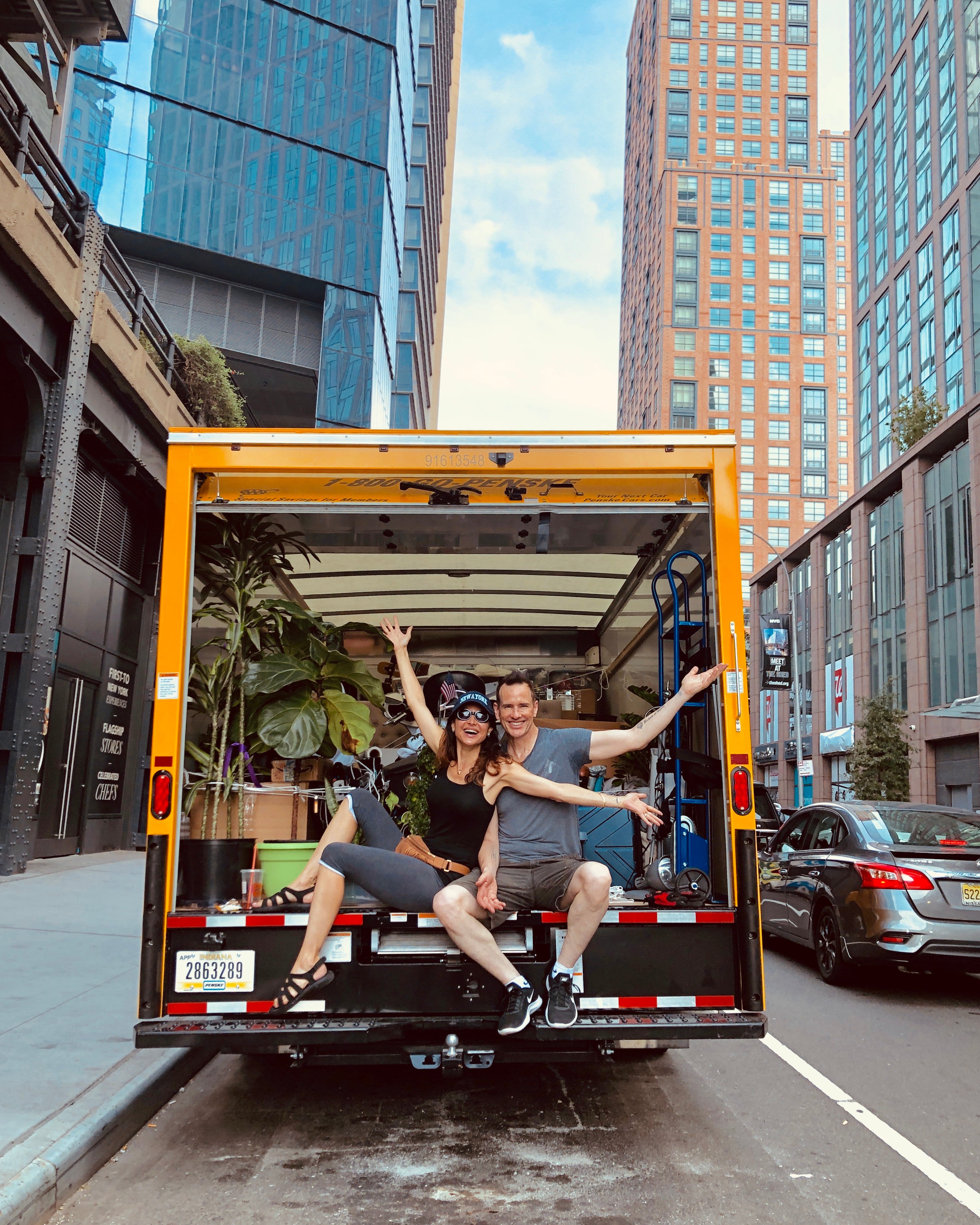 A man and a woman pose happily inside the back of a yellow delivery truck parked on a city street between tall buildings.