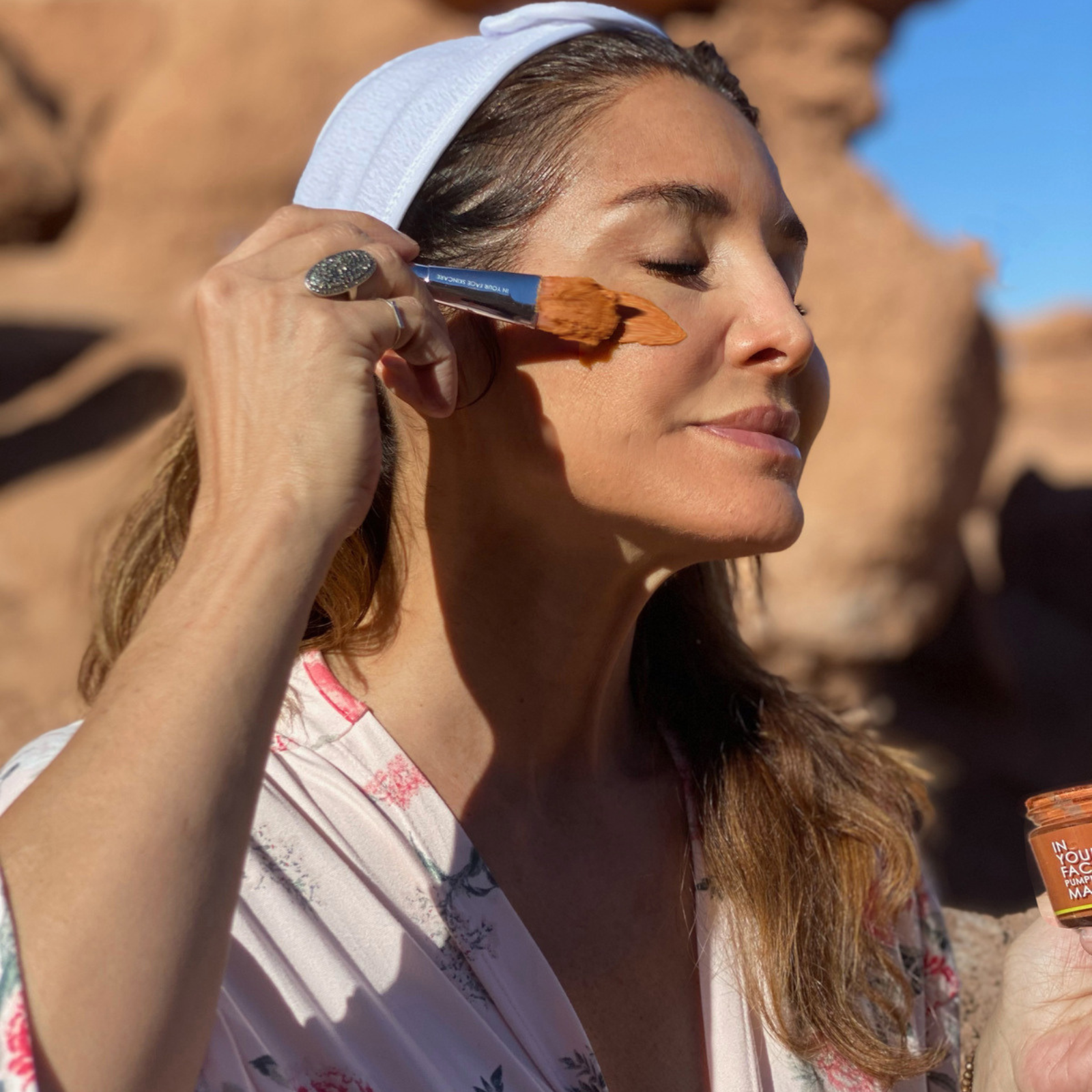 a photo of Denice Duff holding a mask brush to her cheek with some of the PUMPKIN ENZYME MASK on it. She has her eyes closed and a hair wrap on and a flowery robe on. She is in the desert.