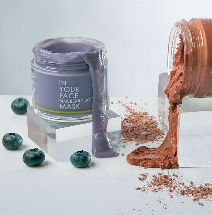 an image of the IN YOUR FACE BLUEBERRY BOOST MASK pouring out with some blueberries around it, and another jar of rust colored face mask coming out