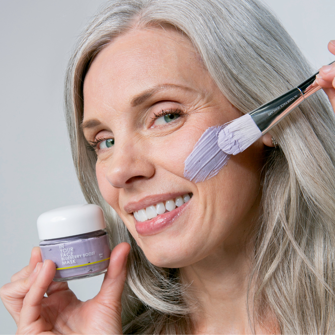 a pretty early 50s woman with long grey hair is shown smiling and applying the BLUEBERRY BOOST MASK to her cheek with an IN YOUR FACE MASK BRUSH.