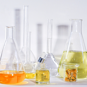 Laboratory glassware with colorful liquids and IN YOUR FACE NOURISHING OIL bottle.