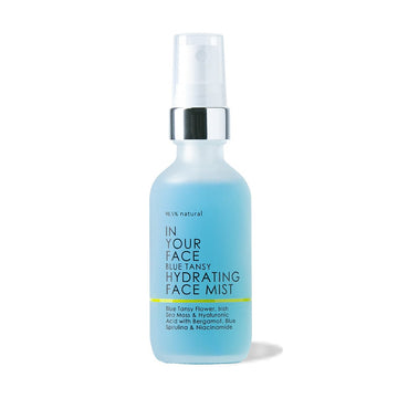 A bottle of the "98.5% natural IN YOUR FACE BLUE TANSY HYDRATING FACE MIST" on a white background. The bottle is clear but slightly frosted and is a bright blue color. The bottle also saying "Blue Tansy Flower, Irish Sea Moss & Hyaluronic Acid with Bergamot, Blue Spirulina and Niacinamide"