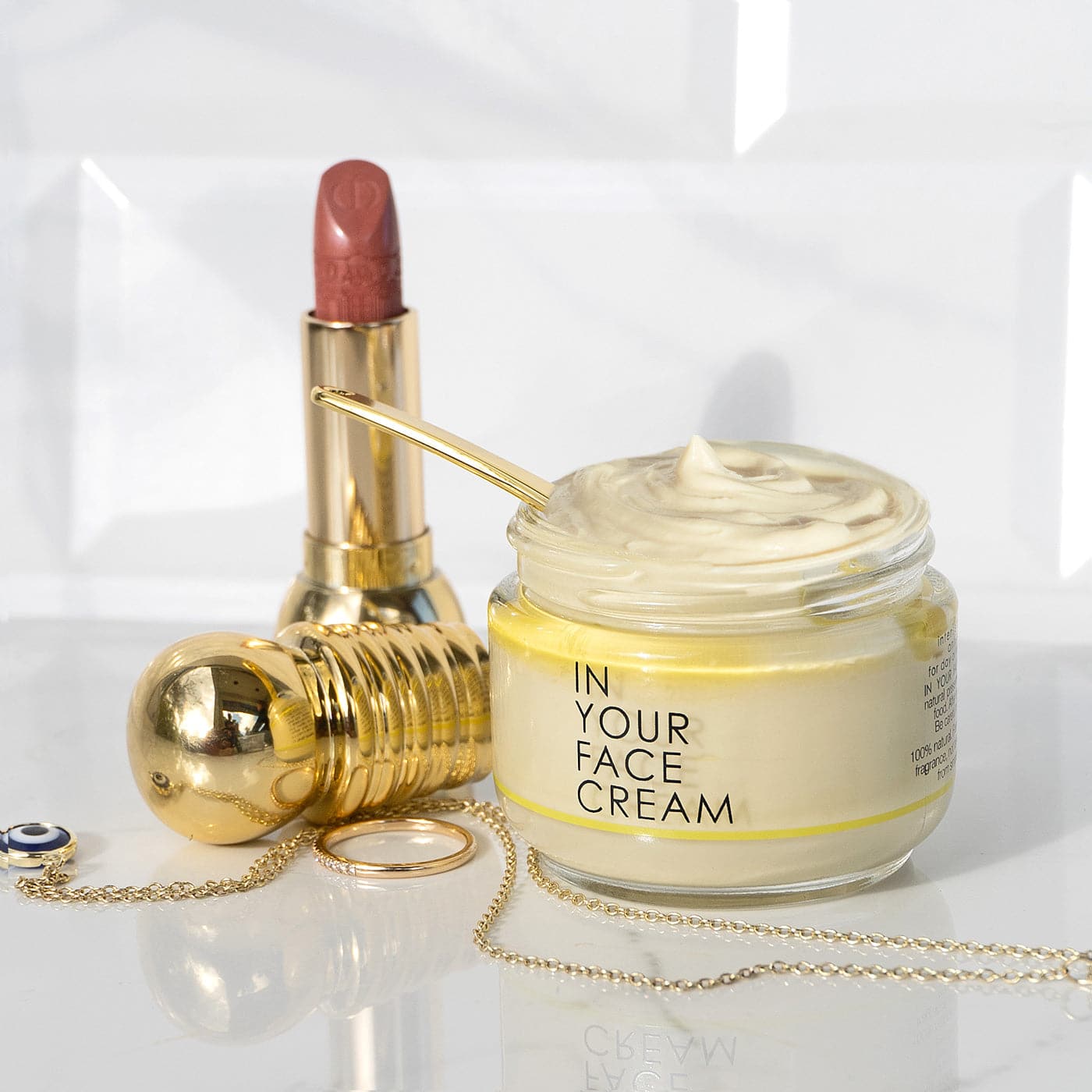 a pretty "lifestyle" image of the IN YOUR FACE CREAM next to a gold tube of mauve lipstick, and a couple pieces of gold jewelry. They are sitting on a white glassy surface. 