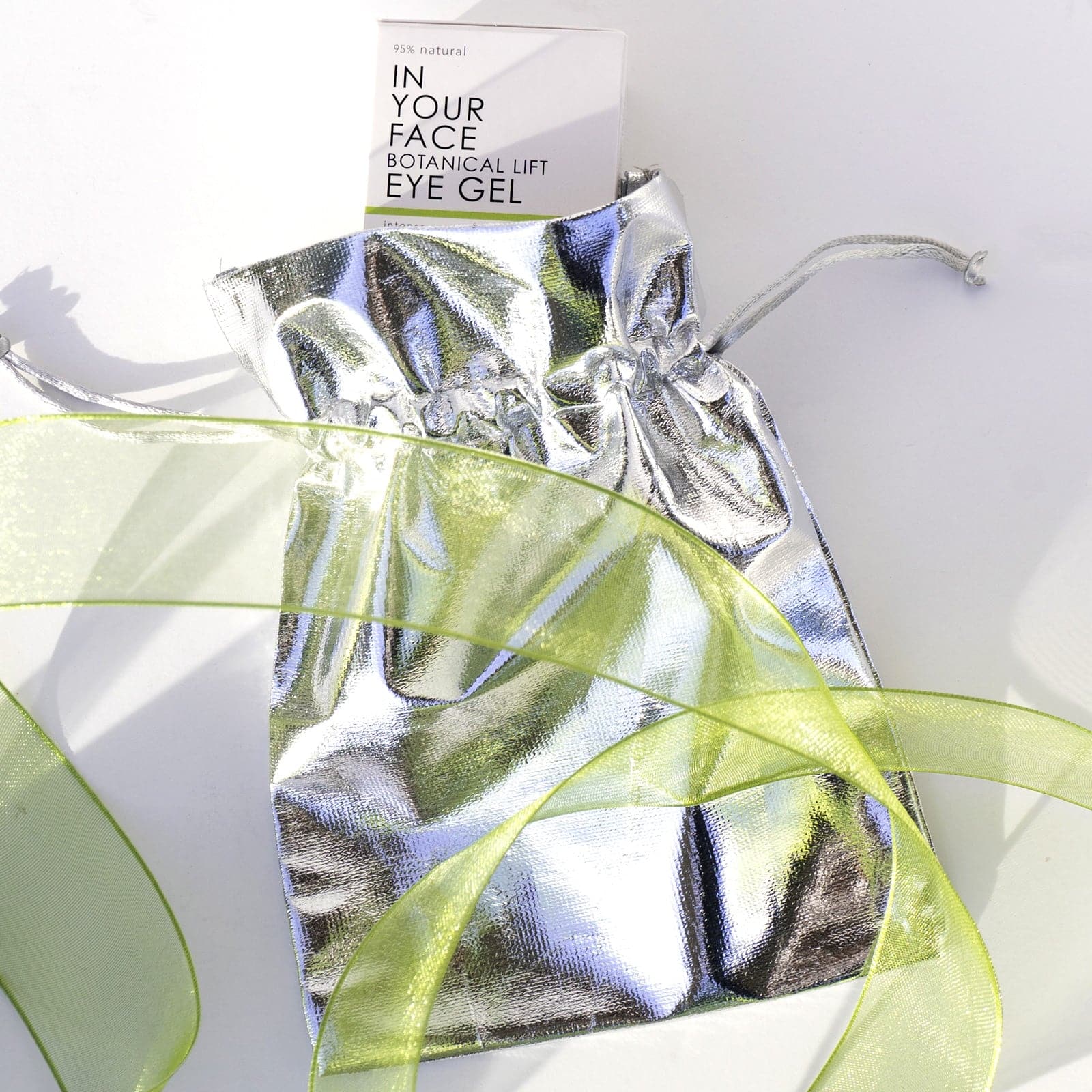 an image of a metallic gift bag with a box of IN YOUR FACE BOTANICAL LIFT EYE GEL popping out. A light green ribbon is wrapped around the bag.