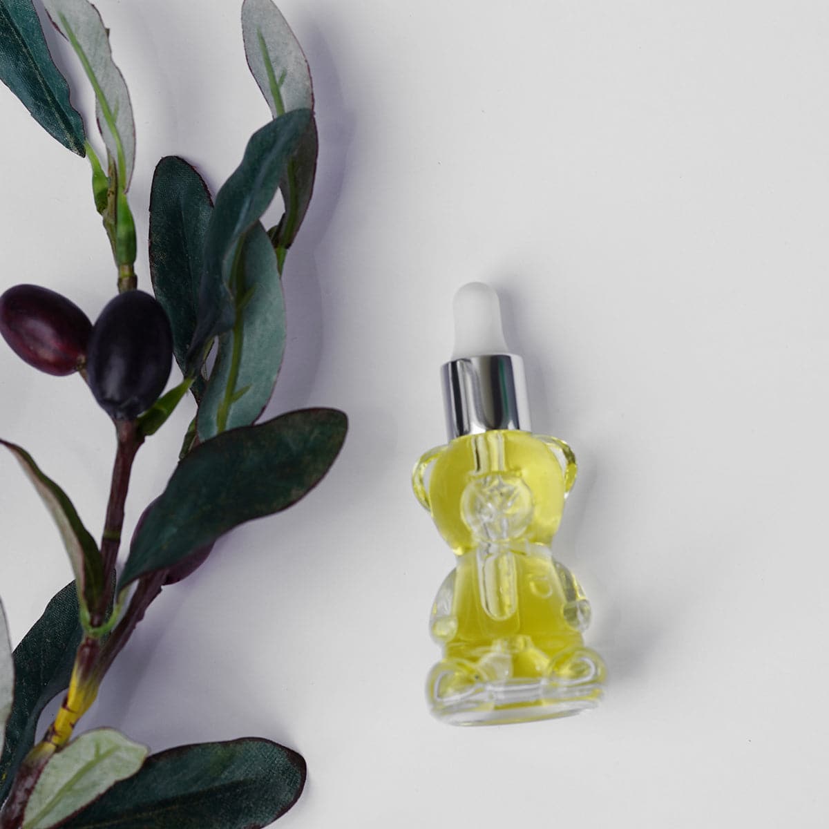 a clean image of the IN YOUR FACE NOURISHING OIL - BEAR EDITION on a greyish background. The oil is in a clear Bear-shaped bottle with a squeezable dropper inside. The oil is a beautiful clear golden color. It's next to an olive branch.