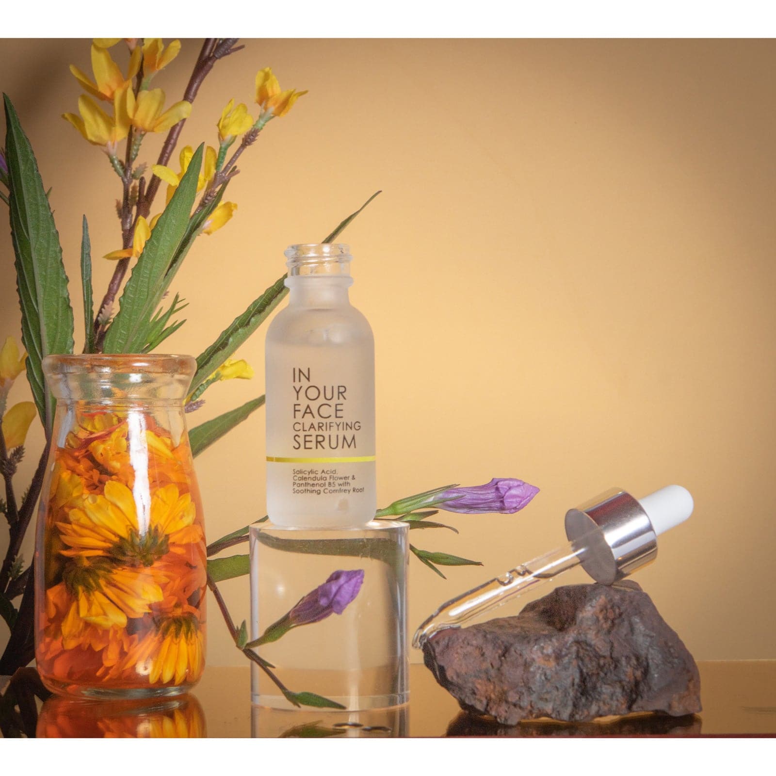 a bottle of the IN YOUR FACE CLARIFYING SERUM on an acrylic cylinder, surrounded by a jar of caledula flowers and various other botanicals. The pipette for the bottle is resting on a rock on the right of the bottle.