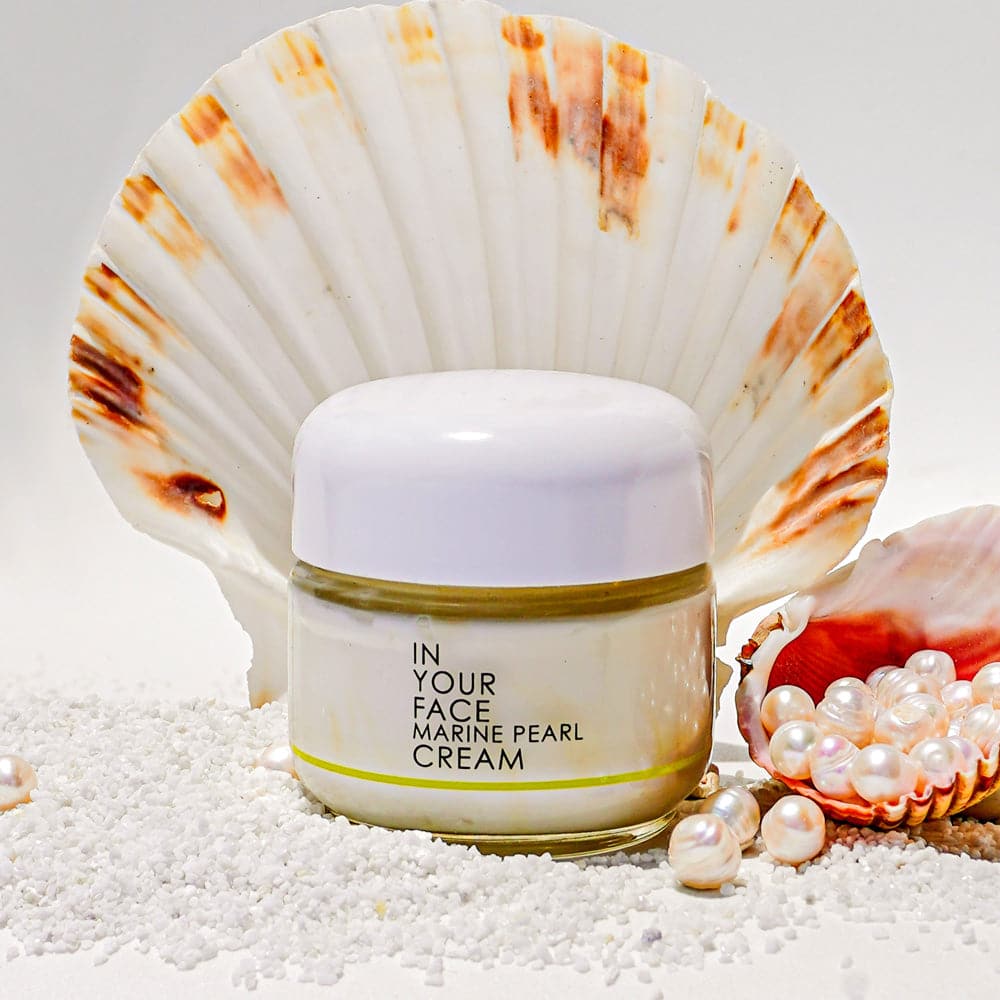 a jar of IN YOUR FACE MARINE PEARL CREAM with a white and brown shell behind it, and both sitting on white sand. Also a white background behind it. And next to both are a shell with about 10 white pearls inside it, spilling out.